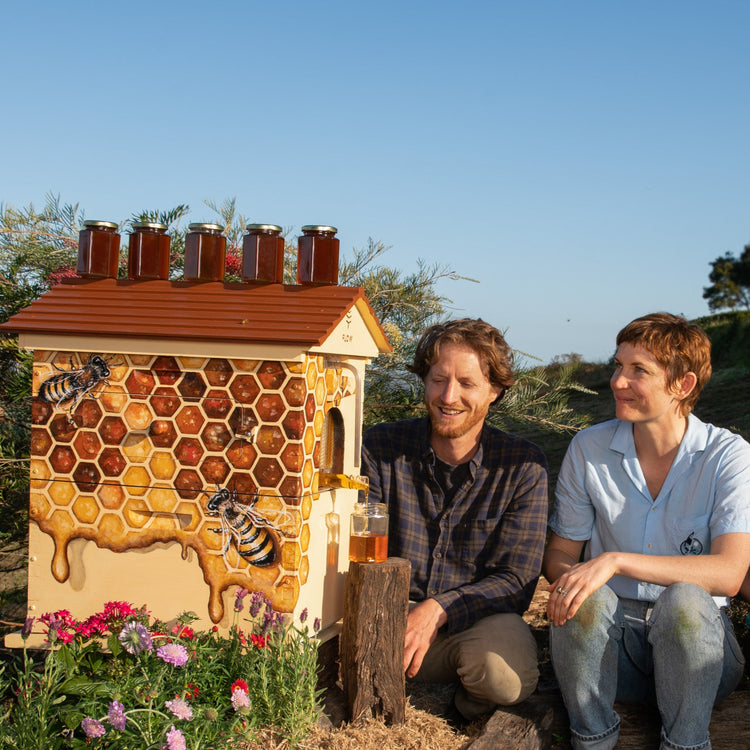 Two people sitting in front of a beehive with honeycomb design, surrounded by flowers and greenery.