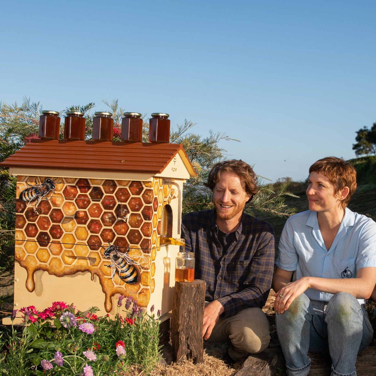 Two people sitting in front of a beehive with honeycomb design, surrounded by flowers and greenery.